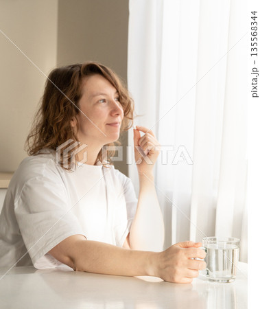 Woman takes pills with a glass of water at home in soft natural light. Daily medication and healthcare concepts 135568344