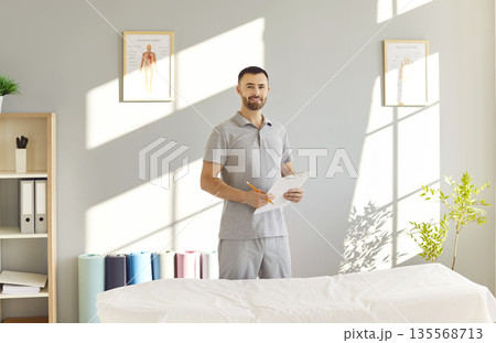 Male physical therapist standing in rehabilitation hospital and looking cheerful at camera. Male physical therapist standing in rehabilitation hospital and looking cheerful at camera. 135568713