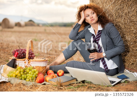 Female office manager with glass of wine and laptop on picnic in field Female office manager with glass of wine and laptop on picnic in field 135572326