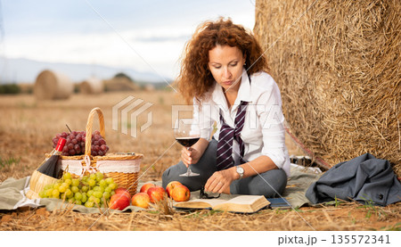 Businesswoman relaxing with wine and book on private picnic near haystack Businesswoman relaxing with wine and book on private picnic near haystack 135572341