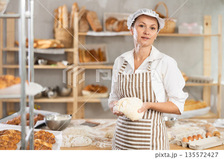 Portrait of positive young woman baker posing with dough in hand in bakery kitchen Portrait of positive young woman baker posing with dough in hand in bakery kitchen 135572427