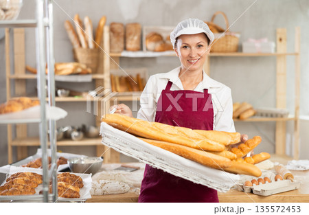 Female baker presenting wicker tray of fresh baguettes Female baker presenting wicker tray of fresh baguettes 135572453