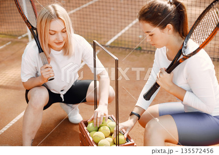 Female friends with racquets and box of balls on the tennis court. Summer, outside court. 135572456
