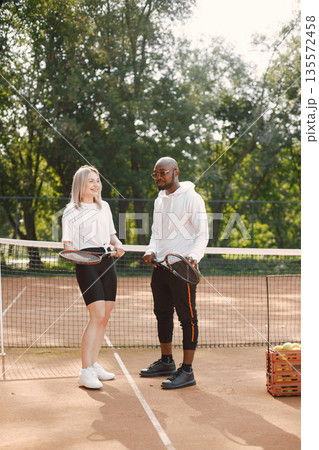 Sumer season. Young black guy with racket and girl having conversation at playground. Tennis, hobbies. Sumer season. Young black guy with racket and girl having conversation at playground. Tennis, hobbies. 135572458