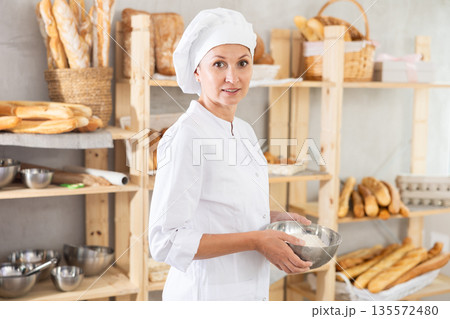 Smiling female baker holding bowl with flour in bakery Smiling female baker holding bowl with flour in bakery 135572480