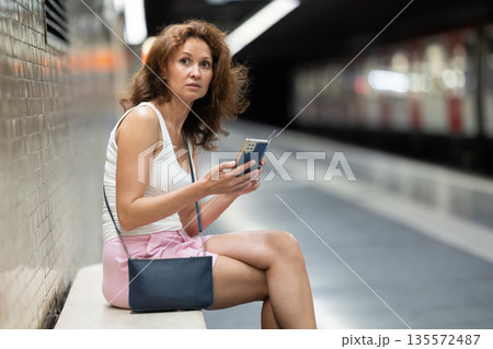 Woman looking for subway train schedule on smartphone while sitting on bench at underground metro station Woman looking for subway train schedule on smartphone while sitting on bench at underground metro station 135572487