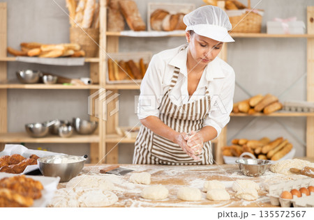 Woman baker forming dough pieces on floured table in bakery Woman baker forming dough pieces on floured table in bakery 135572567