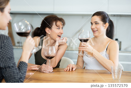 Three happy middle-aged women sitting at table with a glass of wine while having lively conversation Three happy middle-aged women sitting at table with a glass of wine while having lively conversation 135572570