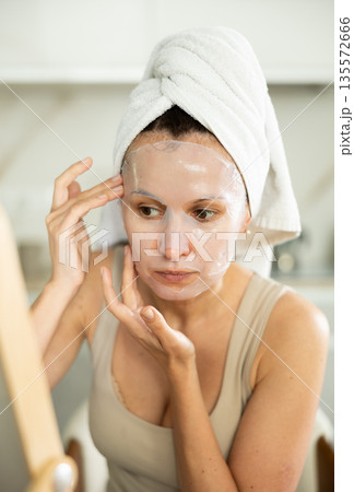 Middle-aged woman applying tissue mask on her facial skin sitting in front of the mirror 135572666