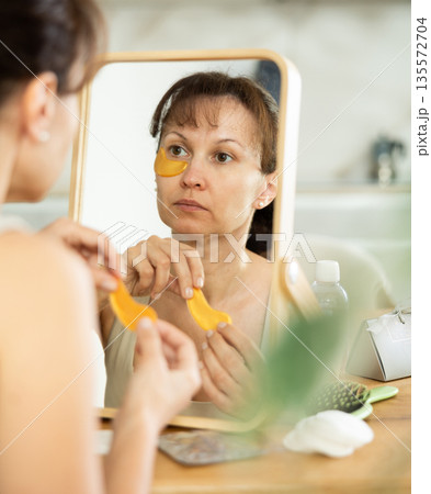 Middle-aged woman applying eye patch mask on her face sitting in front of the mirror 135572704