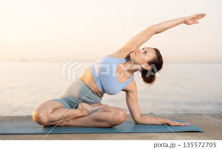 Happy young experienced woman fitness teacher performs yoga parshvottanasana sitting on beach at morning 135572705