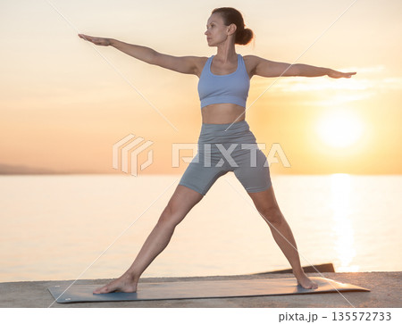 Active experienced woman teacher performs yoga asana on mat on seashore at sunset. Concept of healthy lifestyle Active experienced woman teacher performs yoga asana on mat on seashore at sunset. Concept of healthy lifestyle 135572733