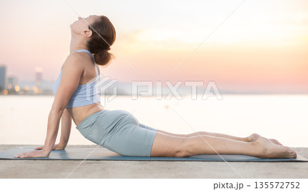 Sportive woman performing cobra pose of yoga during training on beach at sunrise 135572752