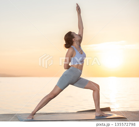 Happy woman coach standing yoga pose and making full body stretching workout on seashore at sunrise 135572798