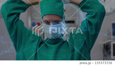 Surgeon in green scrubs and mask securing cap straps in operating room, with digital overlay 135573726