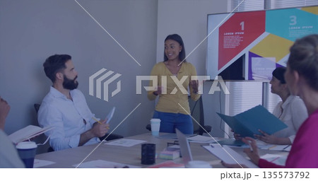 Presenting professional in mustard blouse gesturing toward display screen at boardroom with laptops 135573792