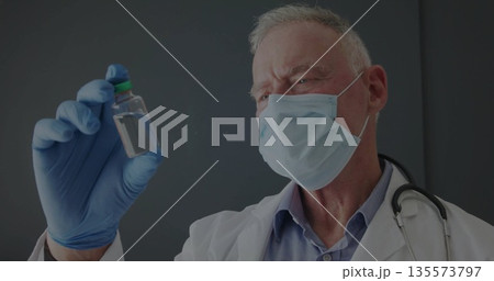 Senior male doctor in lab coat inspecting clear vial at clinic, with stethoscope gloves and mask 135573797