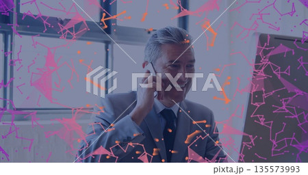 Talking man in suit viewing monitor and using smartphone at desk by window, with network graphics 135573993