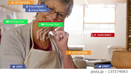 Smiling senior craftsperson touching cheek at studio table, showing ceramic pot and notifications Smiling senior craftsperson touching cheek at studio table, showing ceramic pot and notifications 135574063