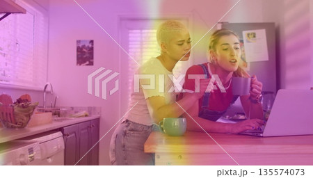 Leaning short-haired woman and her friend examining laptop screen at home kitchen island, with mugs Leaning short-haired woman and her friend examining laptop screen at home kitchen island, with mugs 135574073