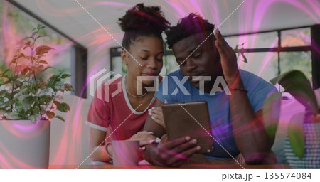 Browsing couple in red and blue shirts viewing tablet at home dining area, with mug, plant 135574084