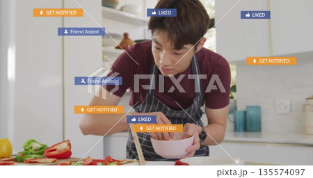 Cooking teenage boy wearing apron holding pink bowl at kitchen counter, with floating notifications 135574097
