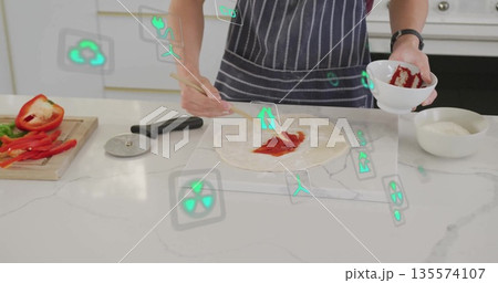 Teenage boy wearing apron smearing tomato sauce on dough in home kitchen, with spoon, eco icons Teenage boy wearing apron smearing tomato sauce on dough in home kitchen, with spoon, eco icons 135574107
