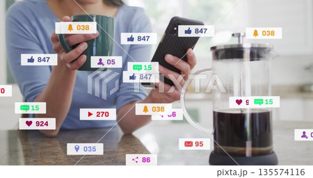 Korean woman holding phone and mug at kitchen counter, with French press, social media icons 135574116