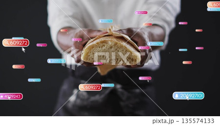 Displaying baker wearing apron holding halved bread in brown paper in studio, with digital tags 135574133