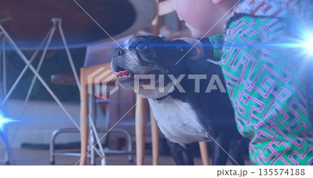 Boy wearing green geometric top kneeling on floor in dining area, petting black-and-white dog 135574188