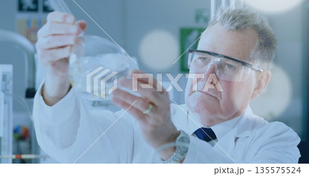 Scientist wearing lab coat and goggles examining glass flask at eye level in lab, with monitor Scientist wearing lab coat and goggles examining glass flask at eye level in lab, with monitor 135575524