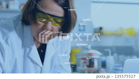 Scientist in lab coat and goggles inspecting beaker at lab bench, with laptop and digital overlays 135575527