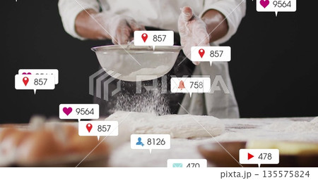 Baker wearing coat and apron shaking flour via fine sieve in kitchen, with notification icons 135575824