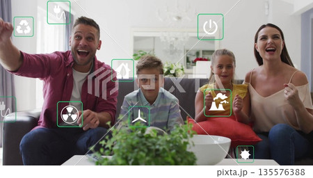 Cheering family raising arms on gray sofa in living room, with floating energy icons 135576388