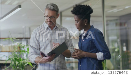 Reviewing black binder man in checkered shirt and woman in denim in modern office, with eyeglasses Reviewing black binder man in checkered shirt and woman in denim in modern office, with eyeglasses 135576448