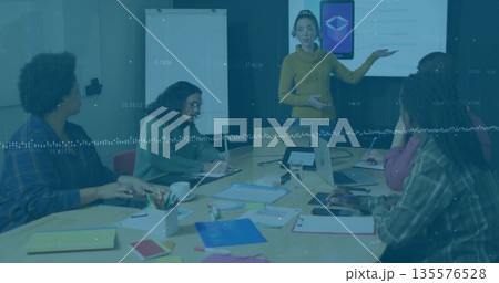 Presenting woman wearing yellow sweater gesturing at display in meeting room, with notebooks, pens 135576528