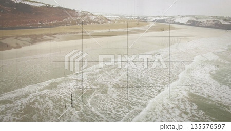 Rolling waves with white foam revealing wet sand on beach, snow-dusted hills, grid overlay hovering 135576597