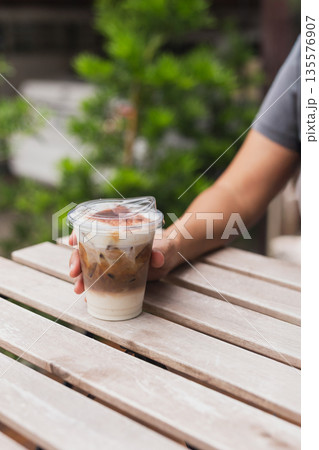 Woman holding iced cappuccino in hand on table outdoors. Woman holding iced cappuccino in hand on table outdoors. 135576907