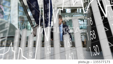 Holding smartphone, businessman in suit riding escalator in glass lobby, with financial charts 135577173