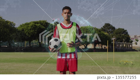 Standing soccer player holding ball wearing neon pinnie near agility poles on training field 135577255