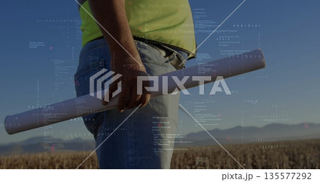 Surveying engineer in high-visibility shirt holding blueprint tube in harvested field, data overlay Surveying engineer in high-visibility shirt holding blueprint tube in harvested field, data overlay 135577292