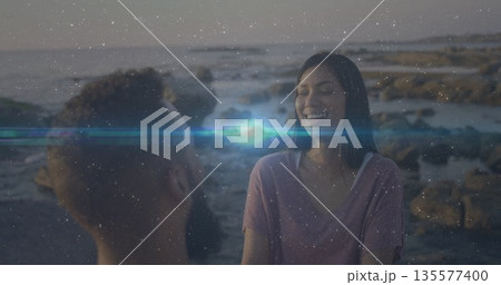 Sitting Hispanic couple wearing purple T-shirt and casual top on rocky shore at sunset, copy space 135577400