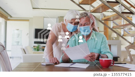 Seniors wearing surgical masks reviewing documents at dining table, with notebook pen coffee mug Seniors wearing surgical masks reviewing documents at dining table, with notebook pen coffee mug 135577415