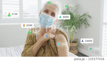 Senior woman pulling down cardigan sleeve revealing bandage at home, with mask, social icons, plant Senior woman pulling down cardigan sleeve revealing bandage at home, with mask, social icons, plant 135577416