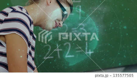 Leaning student with glasses solving math equations at school desk with hologram globe, chalkboard 135577505