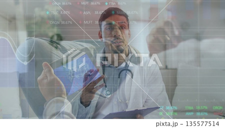 Doctor in coat and stethoscope reviewing tablet at desk, with clipboard, plant and digital overlays 135577514