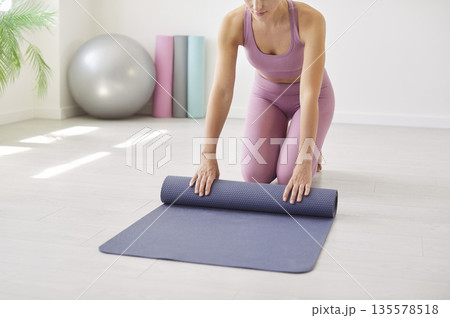 Woman preparing for fitness as she rolls a yoga mat in bright home studio space Woman preparing for fitness as she rolls a yoga mat in bright home studio space 135578518