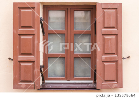 A single window framed by red-brown wooden shutters mounted on a beige wall 135579364