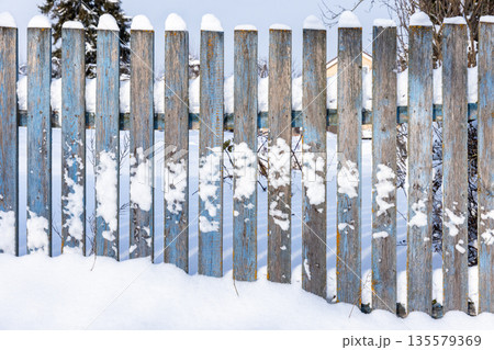A weathered blue wooden fence stands in a snow-filled yard under a crisp winter sky A weathered blue wooden fence stands in a snow-filled yard under a crisp winter sky 135579369