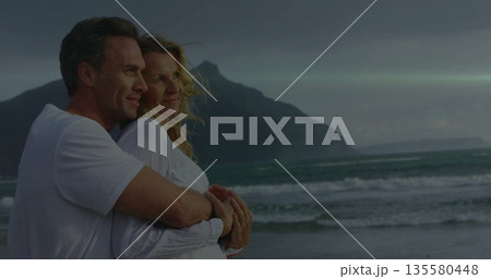 Hugging couple gazing toward horizon at coastal shoreline at dusk, with romantic moment, copy space Hugging couple gazing toward horizon at coastal shoreline at dusk, with romantic moment, copy space 135580448